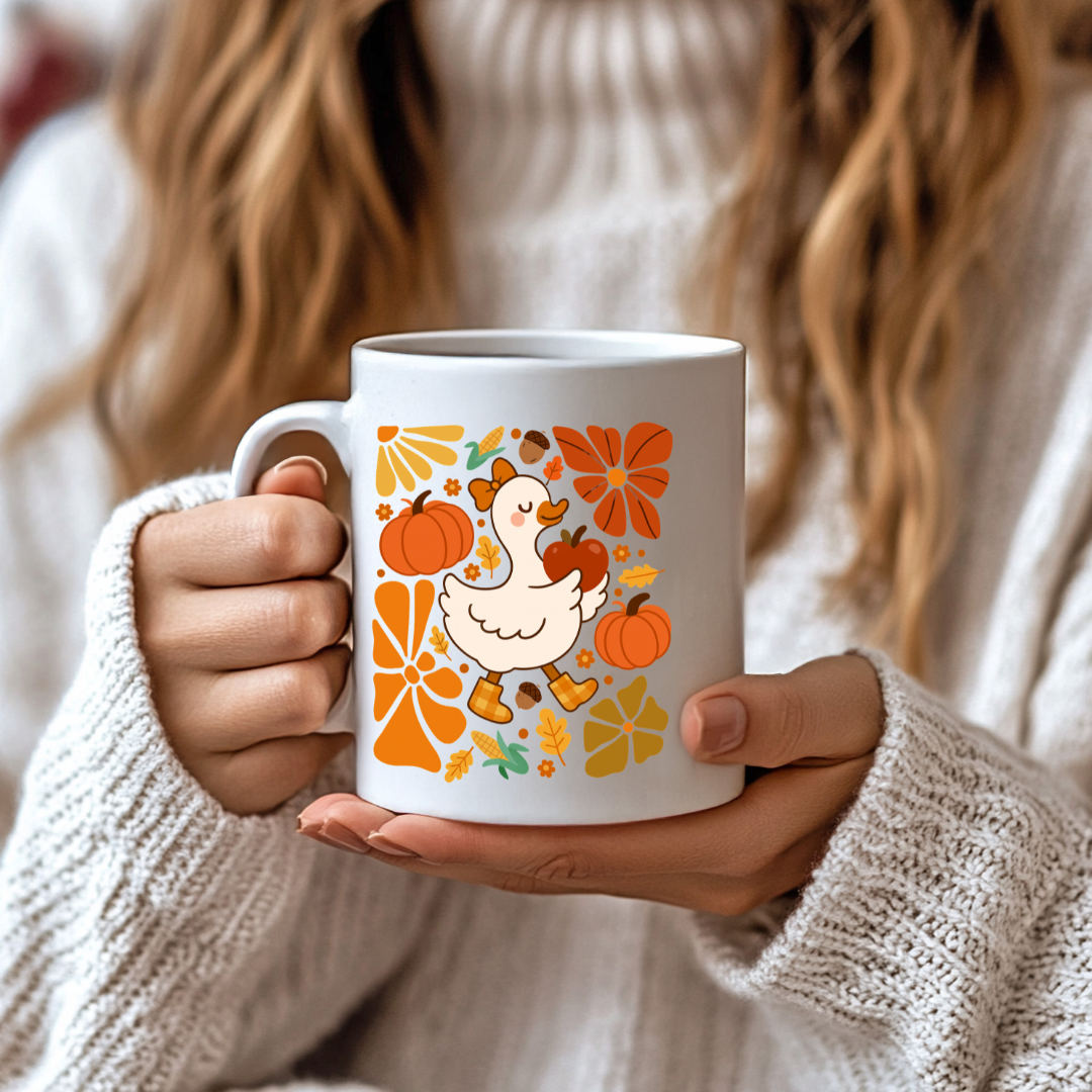 Person holding a white mug with a fall-themed design featuring a duck, pumpkins, and leaves.
