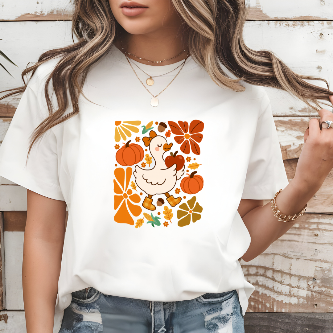 Person wearing a white t-shirt with a graphic design of a duck, pumpkins, and autumn leaves on a wooden background.