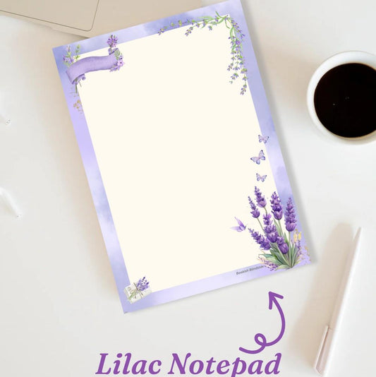 A lilac-colored A6 notepad with floral designs and a pen loop, placed on a desk with a cup of coffee in the background.
