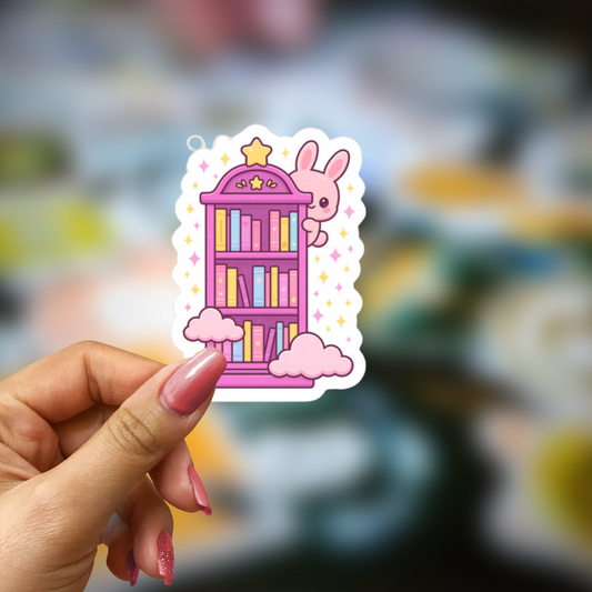 Hand holding a sticker of a pink bookshelf with a bunny character against a blurred background